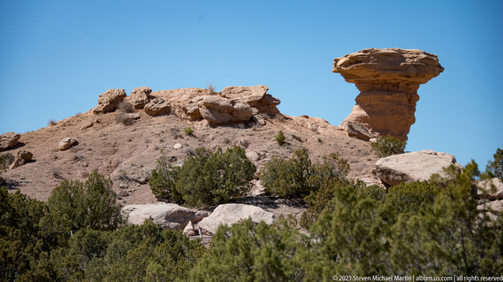 A flat capstone rests on a narrow eroded rock column above shrubs and broken ledges.