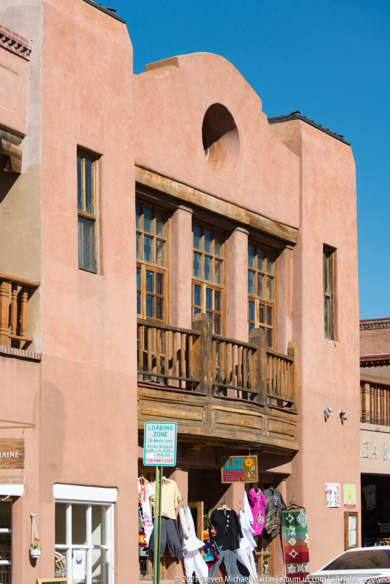 A pink stucco building has timber balconies above ground-floor shops and colorful merchandise.