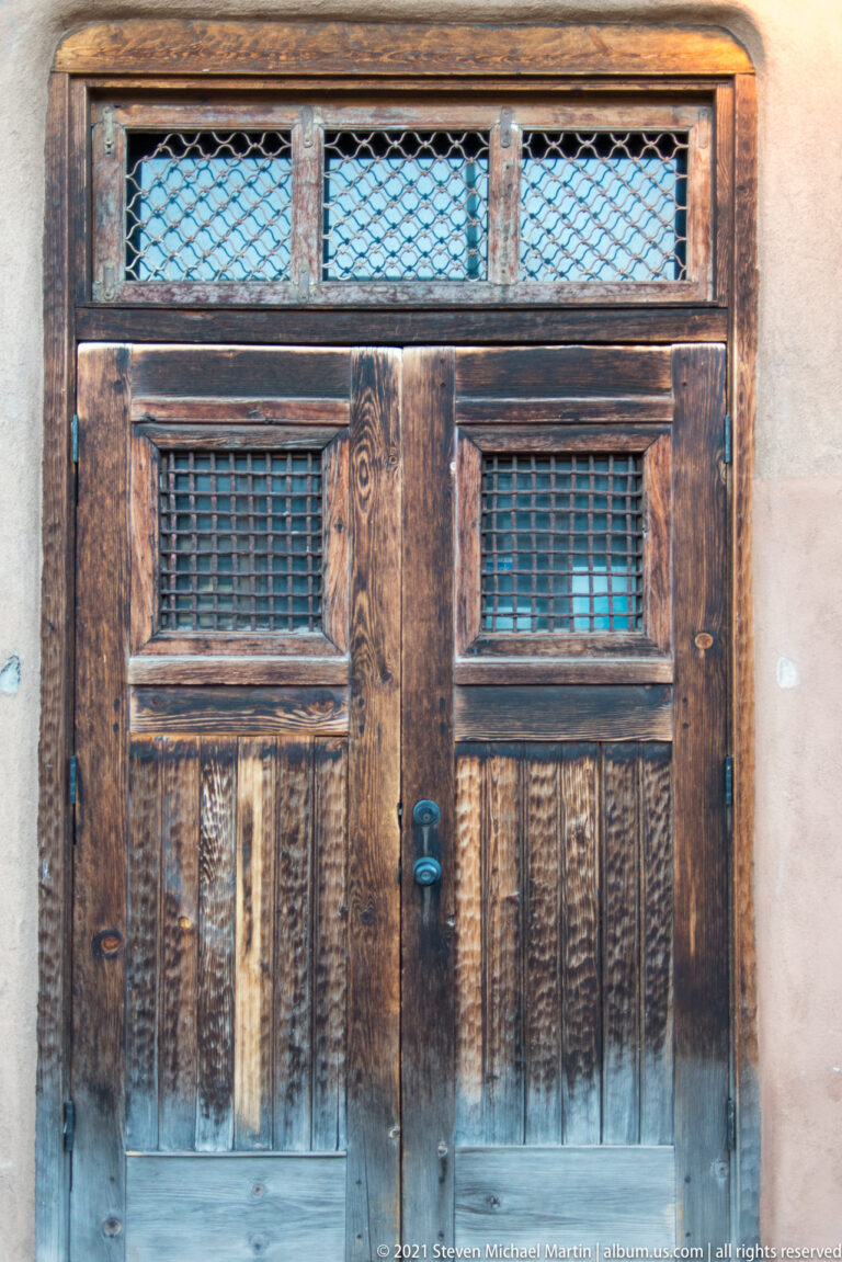 A weathered wooden double door has carved lower panels and small screened openings above.
