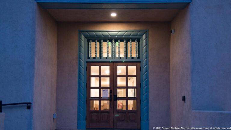 A glass-paneled wooden door sits in a deep stucco recess outlined with blue-green trim.