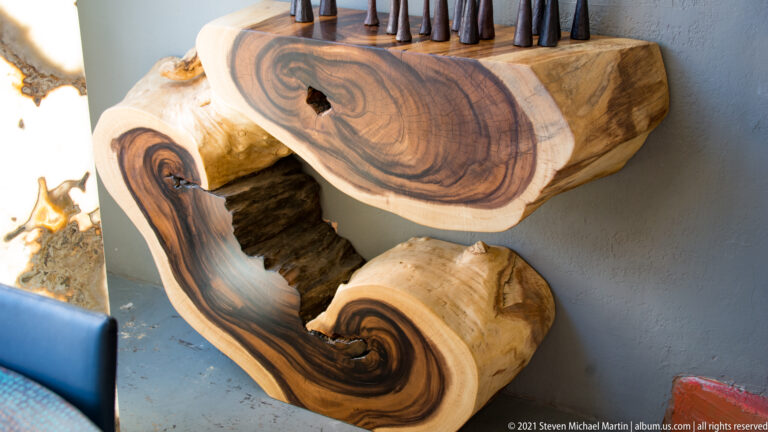 A polished wood sculpture or furniture form has flowing grain, rounded openings, and deep recesses.
