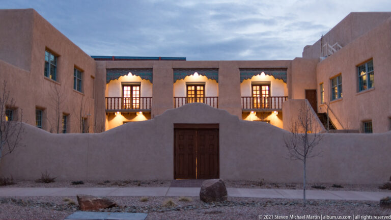 A stucco courtyard facade has a high curved wall, central gate, balconies, and glowing windows.
