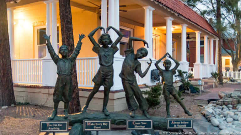 Four bronze child figures pose with raised arms before a white porch and illuminated windows.