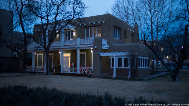 A two-story stucco house with a white balcony and porch is lit from within at dusk.