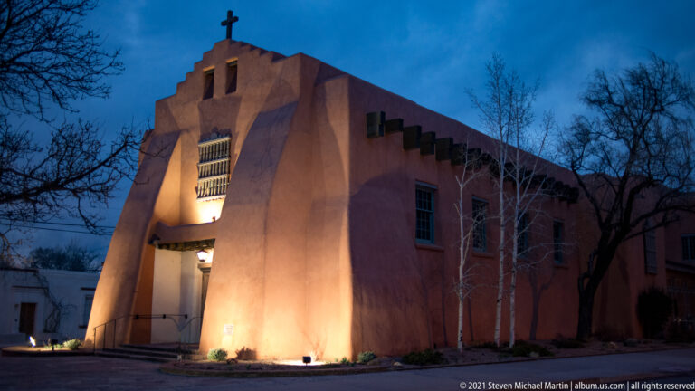 A stepped stucco facade glows at dusk, with a small cross-like form on the roofline.
