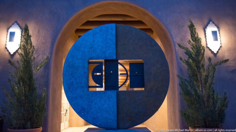 A blue circular door sits inside a rounded stucco arch between two wall lights and evergreens.