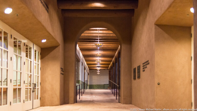 A long hallway with exposed timber beams, pendant lights, glass doors, and wall plaques.
