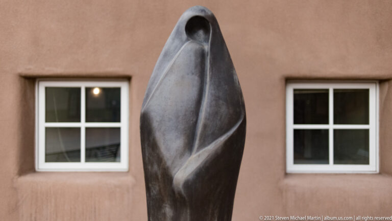 A tall dark abstract sculpture with a small opening stands between two square windows.