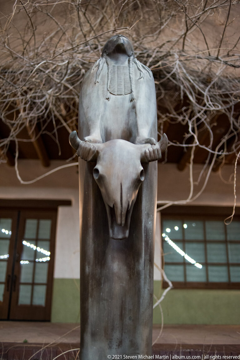 A pale horned animal head projects from a dark vertical sculpture in front of glass doors.