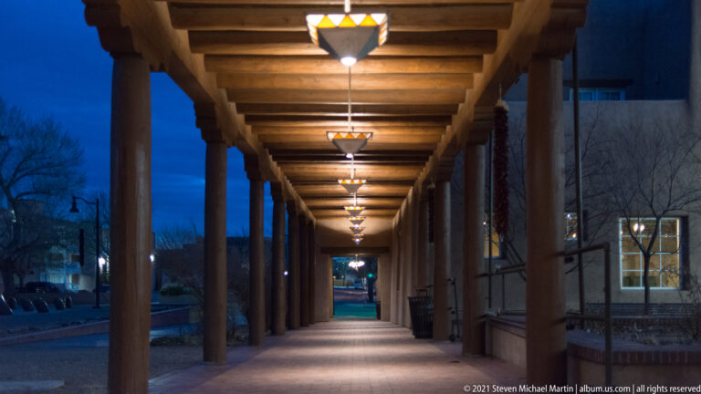 A timber-covered walkway with columns and glowing lanterns recedes into a blue-hour street.