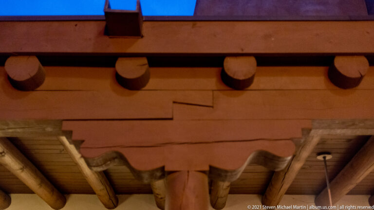 Exposed round roof beams and a carved wooden support sit beneath an adobe-style parapet at dusk.
