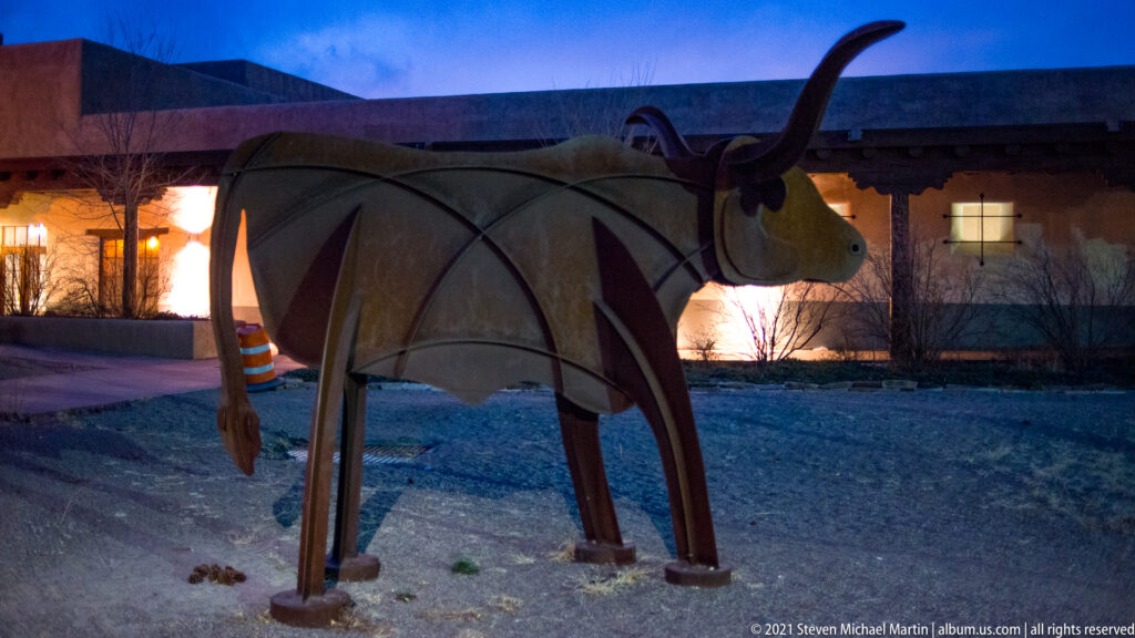 Large angular metal bison with curved horns stands in a courtyard before a warmly lit adobe-style building.
