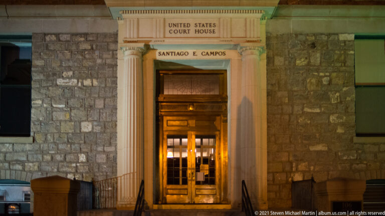 Lit courthouse doorway framed by columns, with signs reading “United States Courthouse” and “Santiago E. Campos.”
