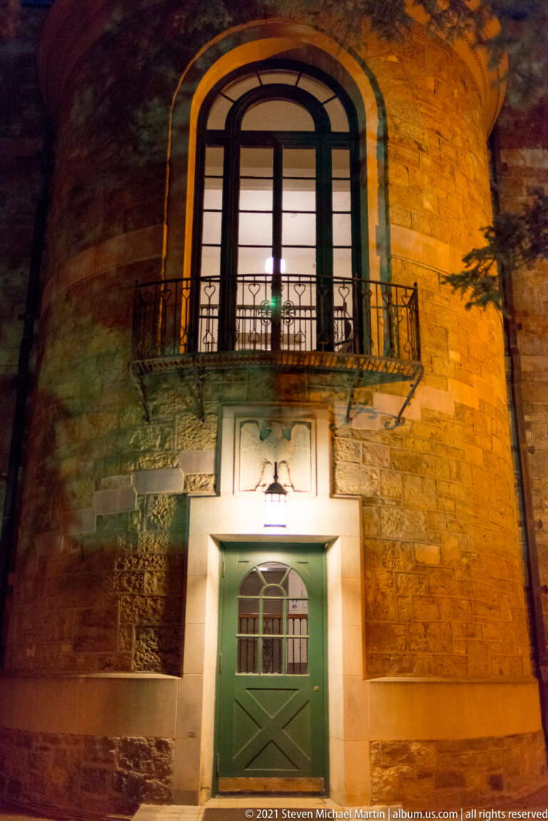 Curved stone façade at night has a green door beneath a small balcony and tall arched window.