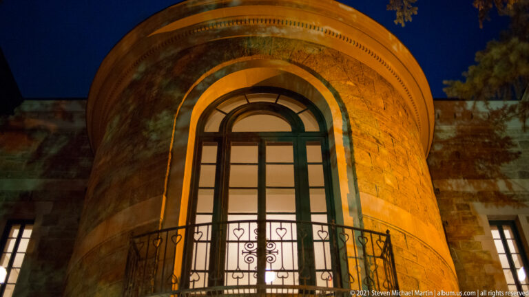 Tall arched window and narrow iron balcony set into a rounded stone tower illuminated at night.