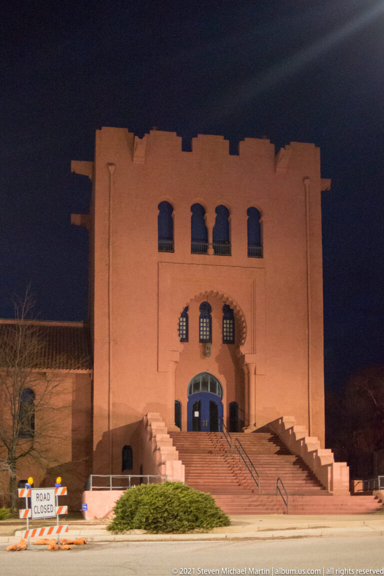 Large stucco building with stepped roofline and central entrance stands above broad stairs at night.