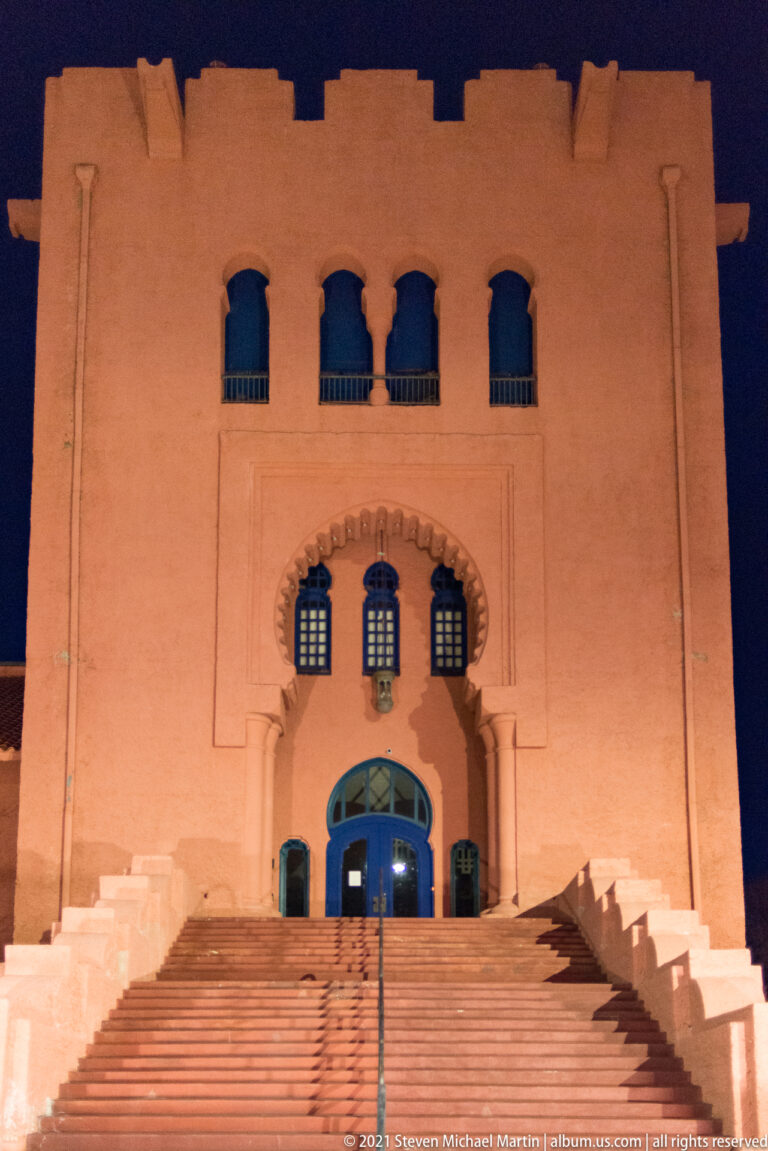 Broad stairs lead to a blue arched doorway below three small windows in a floodlit stucco façade.