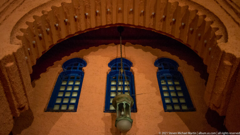 Three blue-framed windows and a hanging lantern sit beneath a deep scalloped stucco arch.