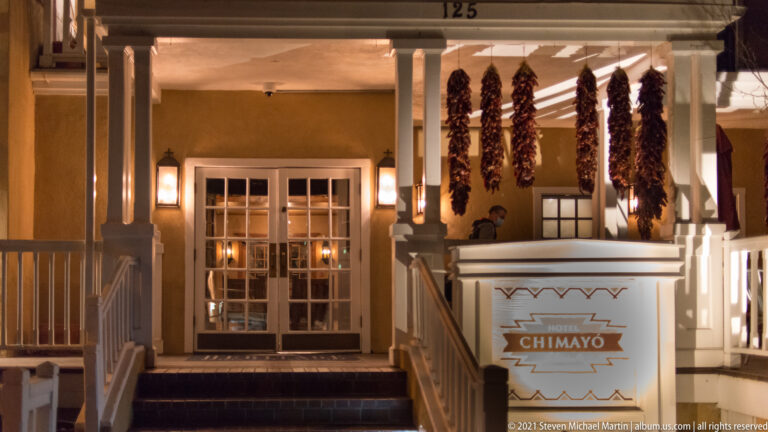 Doorway marked “CHIMAYO” glows at night, with strings of dried red chiles hanging beside it.
