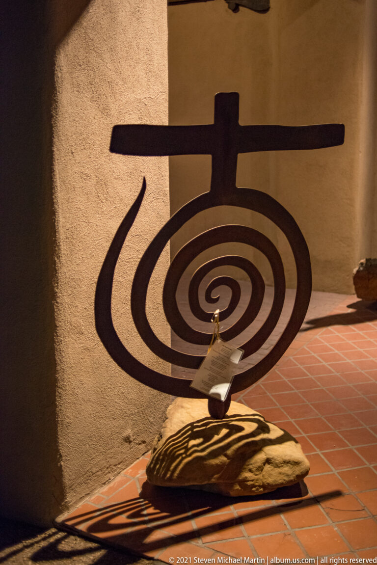 Metal wall sculpture has a crossbar above nested spiral shapes, with a stone form below.
