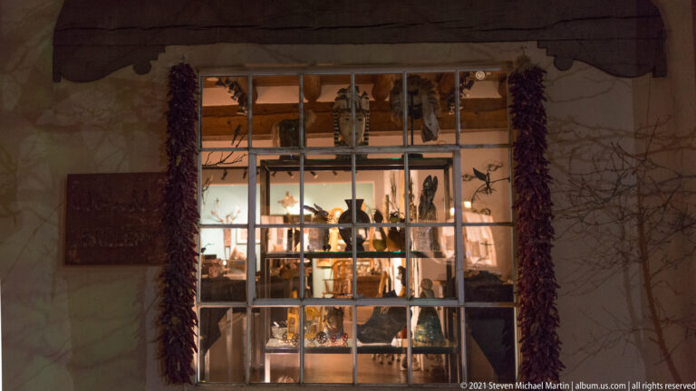 Shop window displays pottery and carved objects behind glass, framed by hanging red chile strings.
