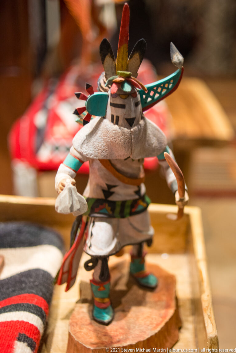 Small figurine in a pale outfit and feathered headgear stands before colorful woven textiles.