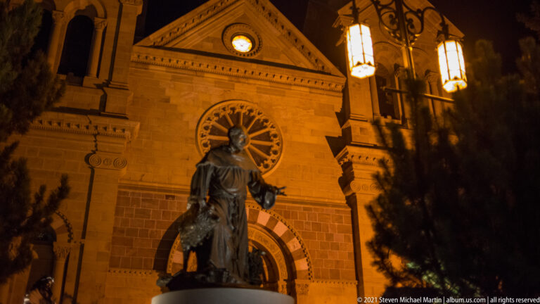 Bronze robed figure viewed from behind stands before the cathedral’s rose window at night.