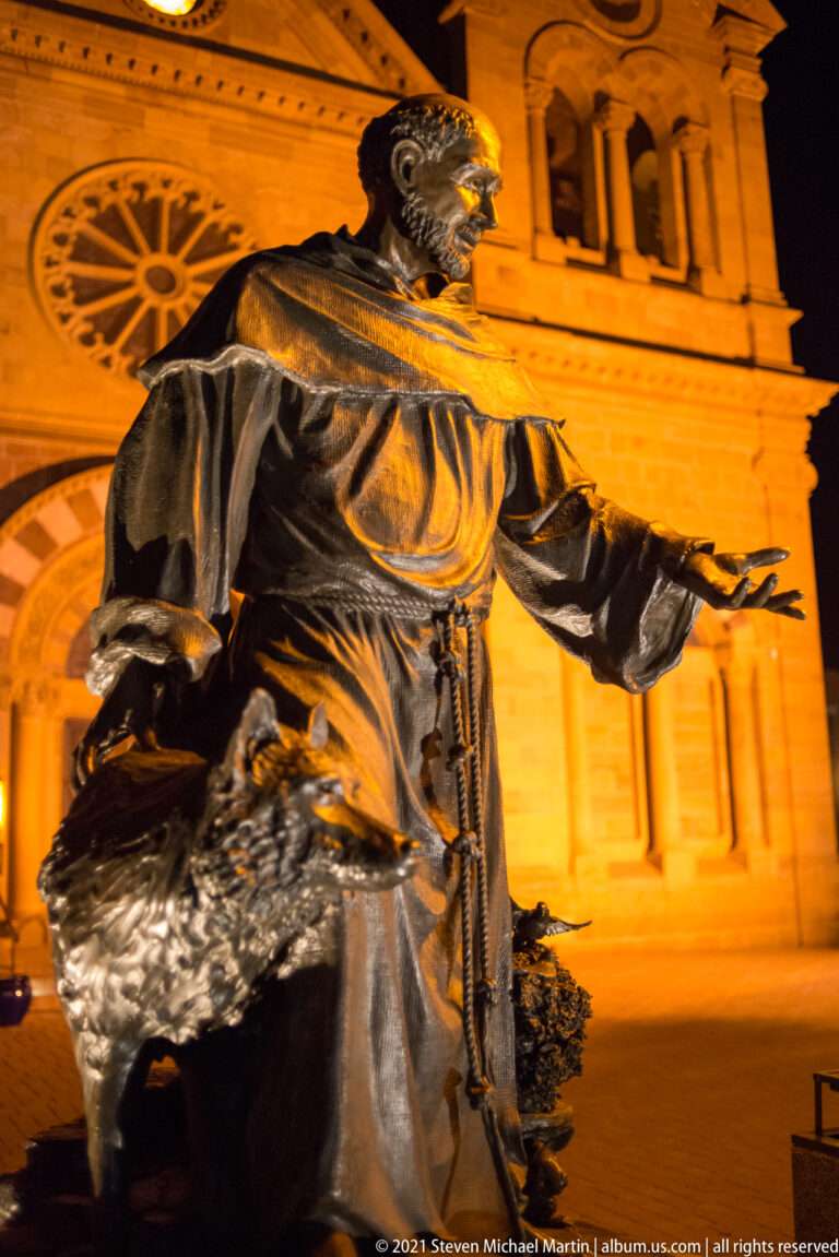 Bearded bronze figure in a robe extends one arm before an illuminated cathedral façade.