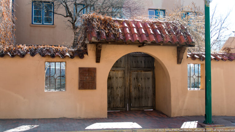 A wooden double door sits beneath a red tiled canopy in a stucco wall with small windows.