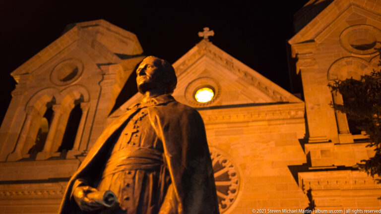 Bearded bronze figure in a belted robe stands before the warmly lit cathedral at night.
