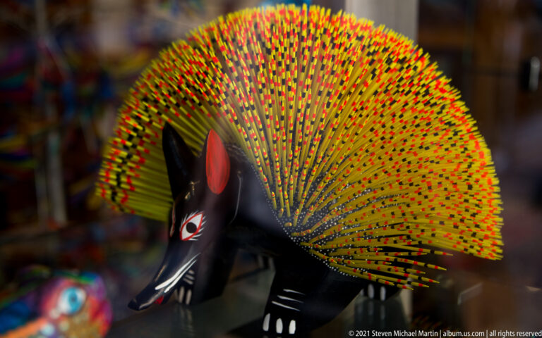 A black animal sculpture with a long snout stands beneath a fan of brightly painted quills.