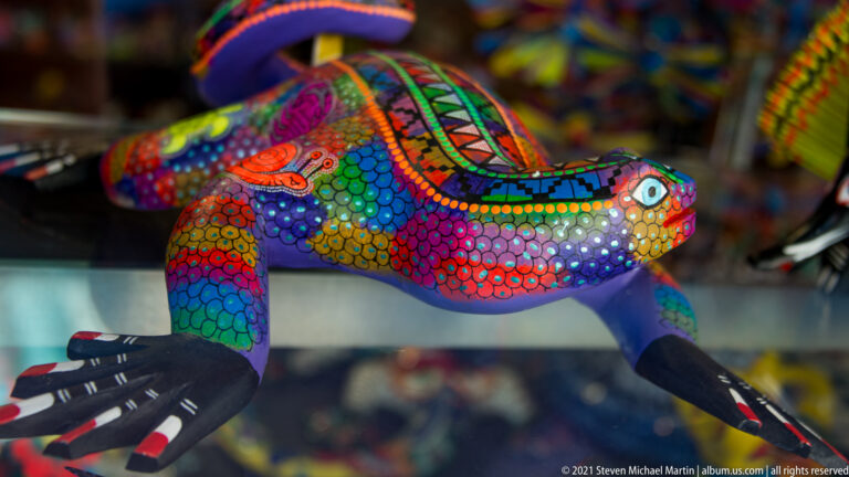 A lizard sculpture with splayed legs is covered in bright multicolored patterns and dots.