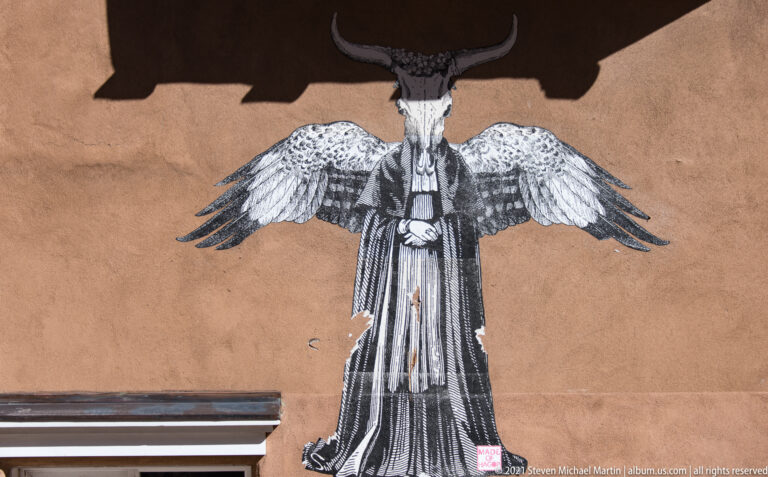A large black-and-white winged figure is painted on a tan stucco wall beneath a roof.
