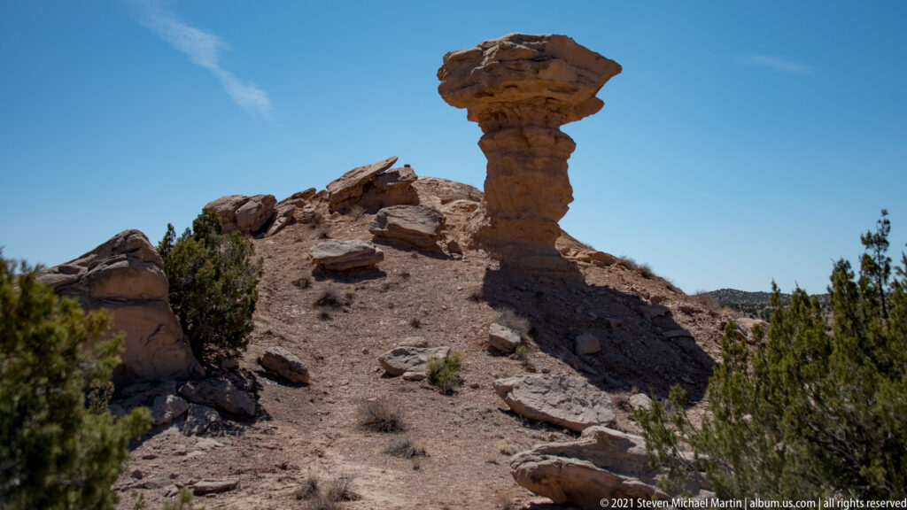 A mushroom-shaped rock formation rises above a broken slope with shrubs and scattered stones.