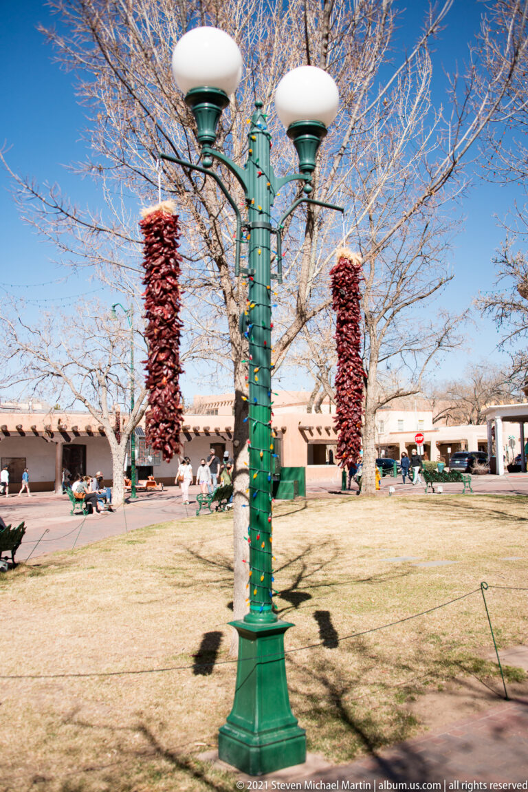A green plaza lamppost is flanked by two hanging strings of dried red chiles.