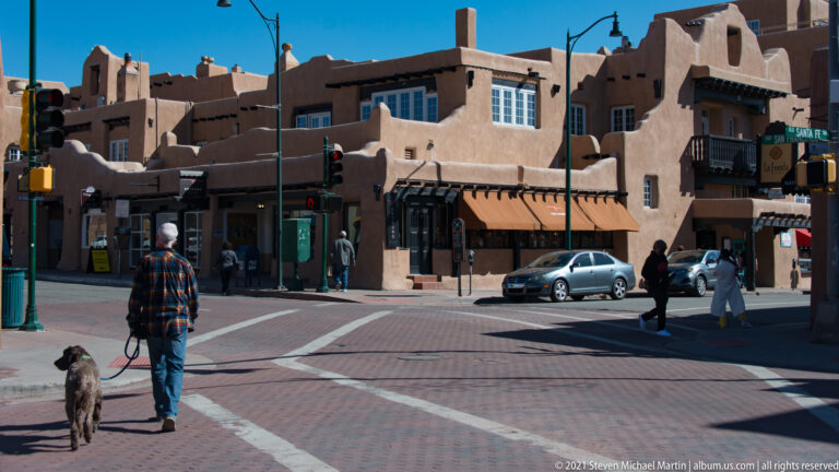 A person walks a dog across an intersection lined with stucco buildings and pedestrians.