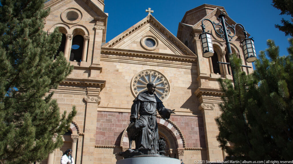A bronze statue stands before a cathedral facade with a rose window, gable, and evergreen trees.