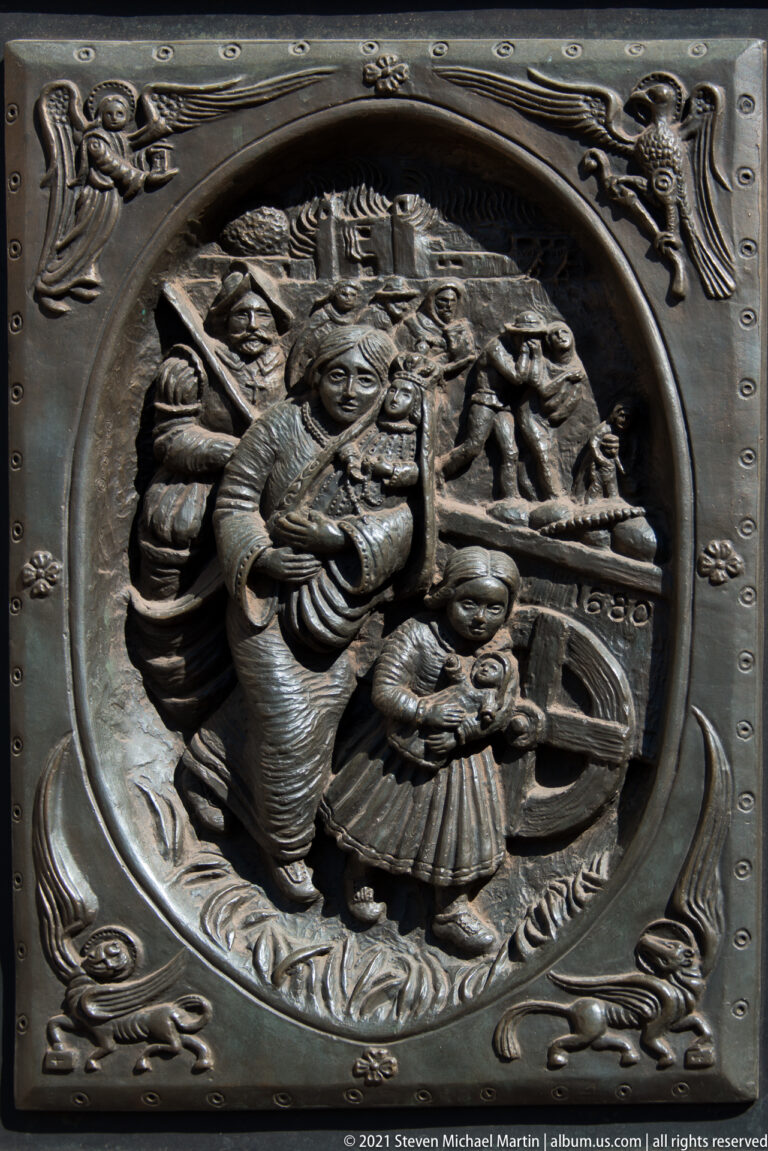 A bronze relief shows robed figures gathered around a seated person within an ornate oval frame.