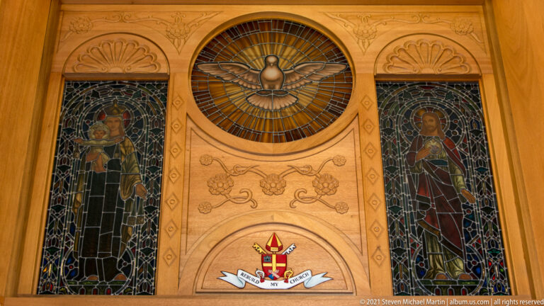 A round stained-glass dove window sits above a coat of arms between two tall saint windows.