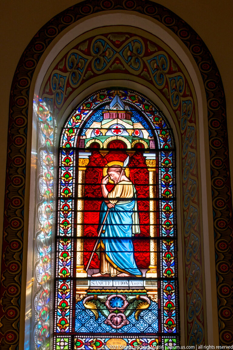 Tall stained-glass window shows a robed figure against red glass, surrounded by a multicolored ornamental border.
