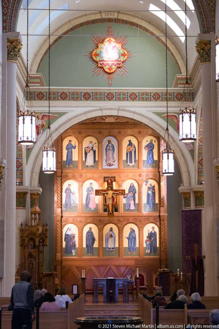 Pews lead toward a painted altar screen with saint figures beneath a decorated arch and hanging lights.