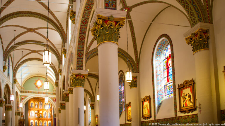 Interior nave recedes through rounded arches, with stained-glass windows, columns, lanterns, and framed religious art.