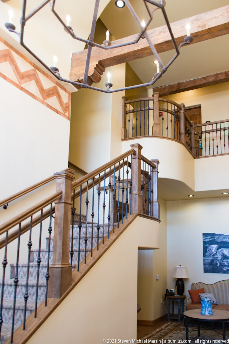 A curved staircase and upper gallery overlook a lobby with seating, framed art, and timber beams.