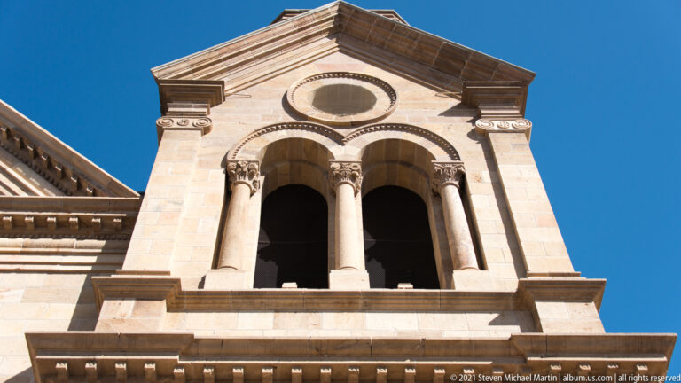 Upward view of a pale stone church tower with a circular opening and paired arched windows against blue sky.