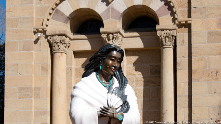 Bronze statue of a woman wearing a white shawl and turquoise jewelry stands before paired stone arches.