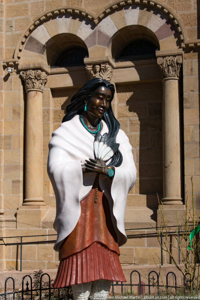 Full-length bronze woman wears a white shawl and red skirt behind a railing before a stone church arcade.