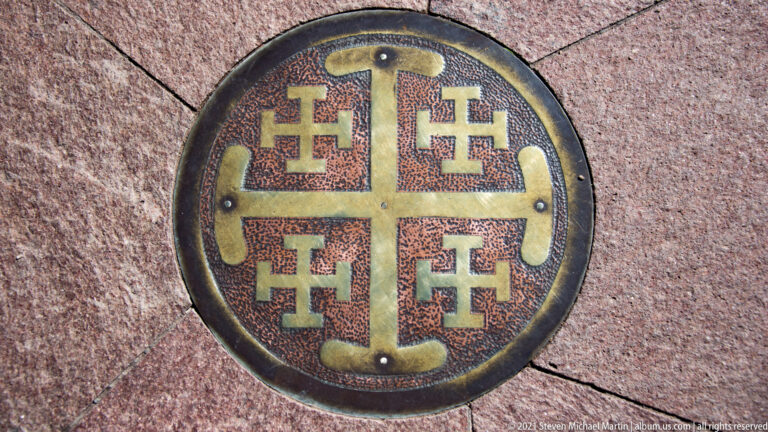 Round wall medallion displays a central cross surrounded by four smaller crosses on a stone background.