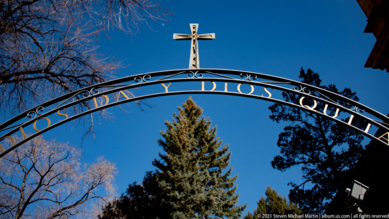 Curved iron entrance arch with partly obscured lettering and a cross stands above a gate.