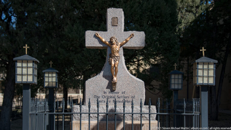 Bronze crucifix rises above an inscribed stone memorial inside a fenced, tree-shaded area.