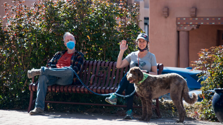 Masked visitor waves from a bench beside another seated person and a large dog.